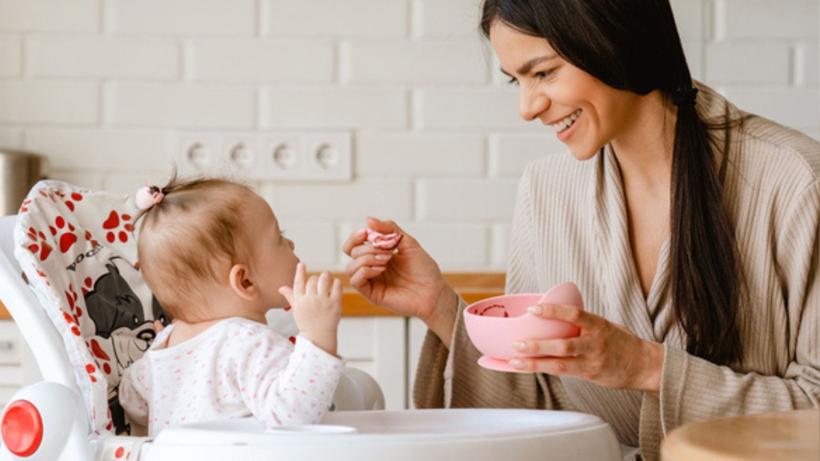 Young brunette mother smiling and feeding her baby in kitchen at home.jpg