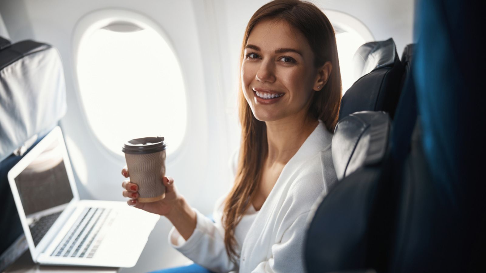 Woman seated at the window side in airplane drinking hot beverage with laptop.jpg